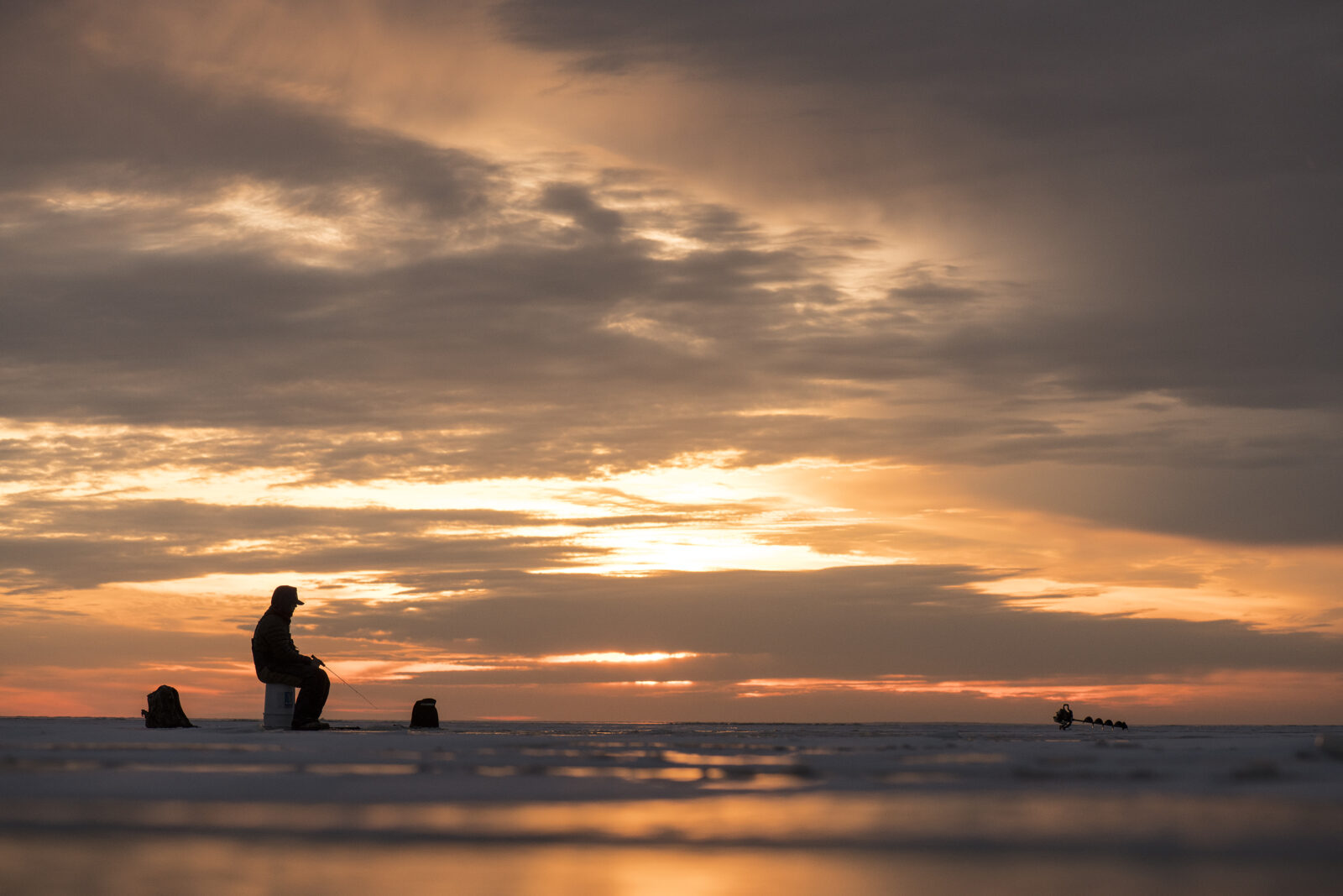 Ice fisherman on a frozen lake with a beautiful sunrise behind him. 