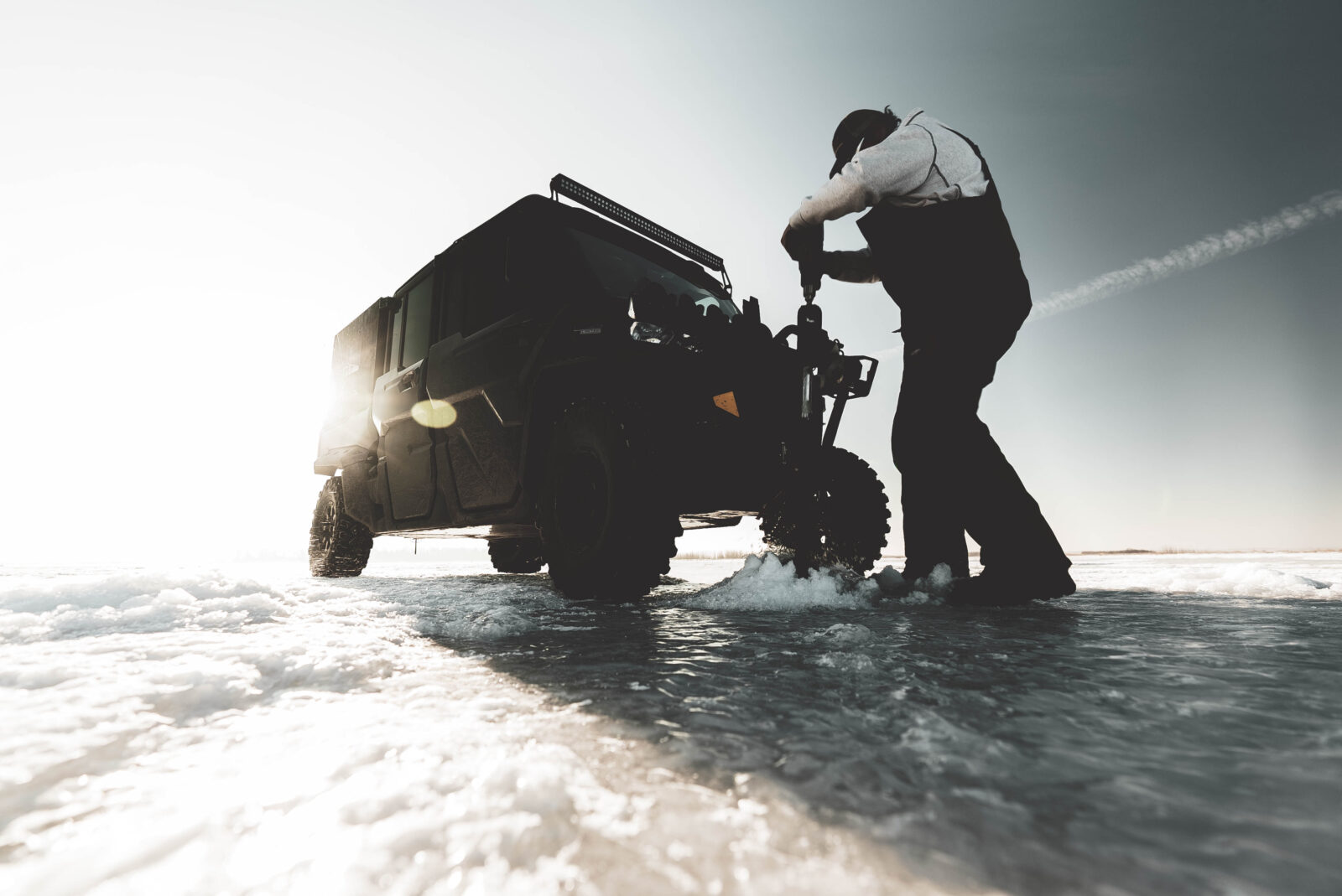 Ice fisherman on a frozen lake using an auger next to his vehicle.